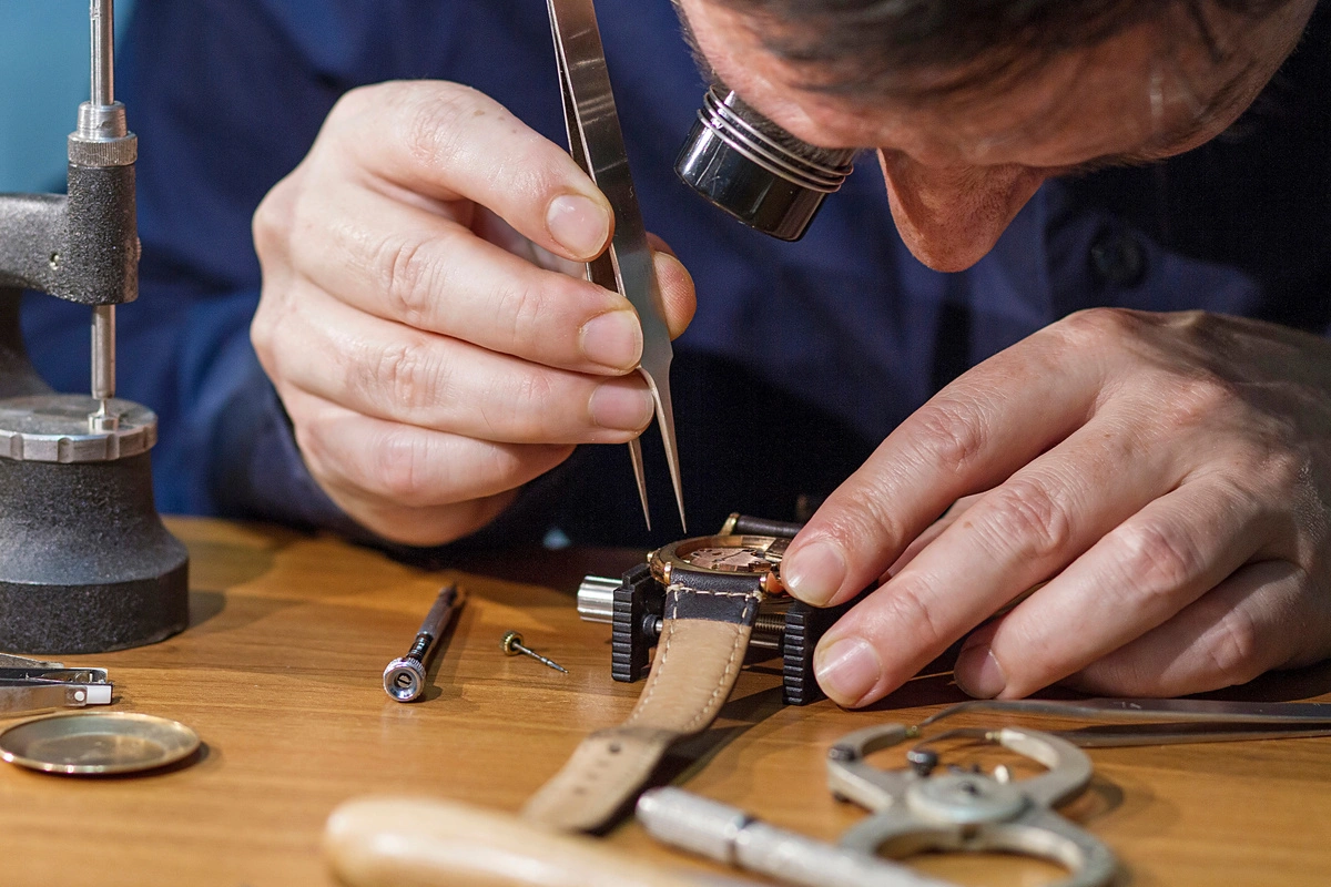 Artisan réparateur de montres travaillant minutieusement avec des pinces sur une montre à bracelet, entouré d'outils horlogers spécialisés.